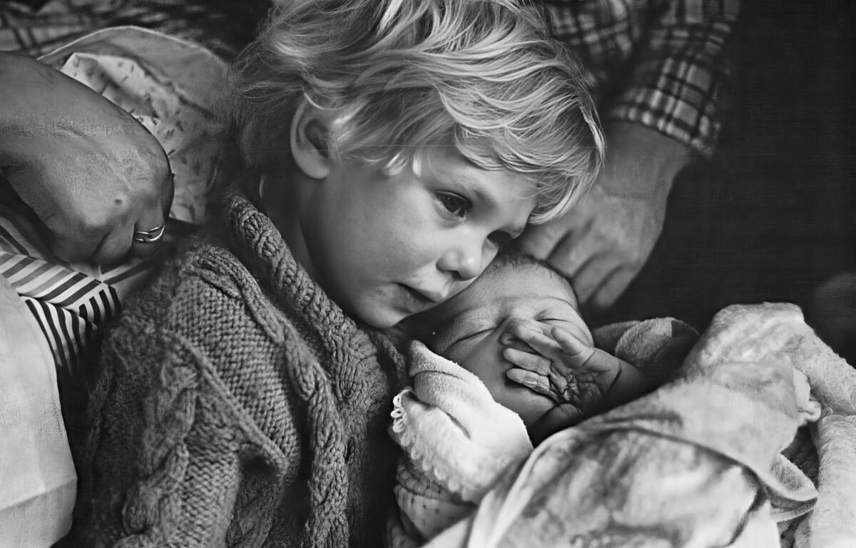 Black and white photo of young boy holding a newborn baby with a hand behind him