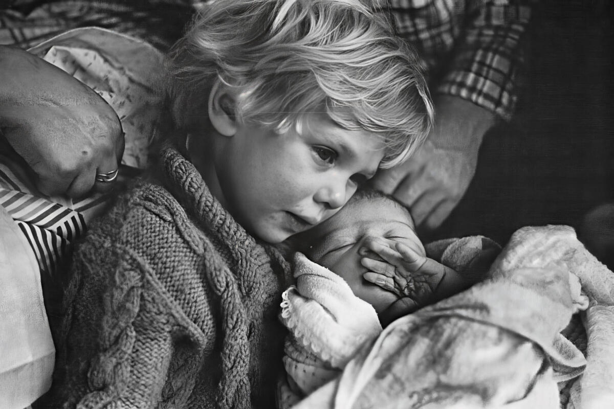 Black and white photo of young boy holding a newborn baby with a hand behind him